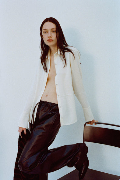 Image of model wearing Maddi Shirt in Merino Wool Twill in Bone and Magnus Pants in Lightweight Leather in Black holding onto a chair against a plain background