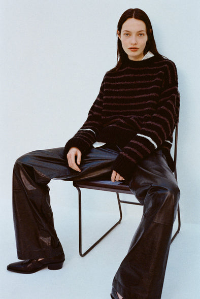 Image of model wearing Leonie Sweater in Stripe Wool in Black multi and Magnus Pants in Lightweight Leather in Black sitting on a chair against a light blue background