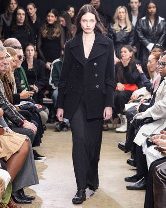 Model walking in Proenza Schouler Fall Winter 2024 Runway show wearing ROWEN PEACOAT IN BLACK ECO DOUBLE FACE WOOL, BELLA DRESS IN BLACK LACQUERED KNIT, and  TRACK SNEAKERS IN BLACK NAPPA/SUEDE