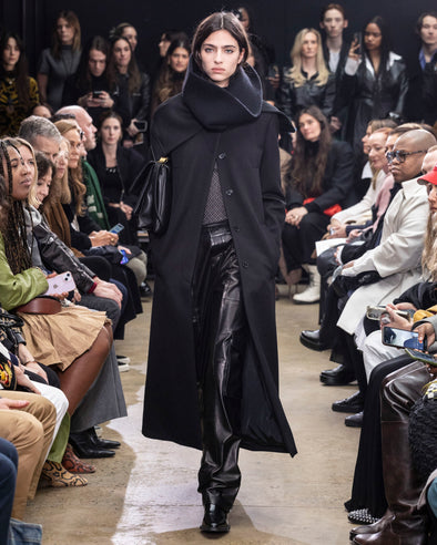 Model Walking In Proenza Schouler Fall Winter 2024 Runway Show Wearing Phoenix Coat In Black Wool Gabardine, Rio Top In Black Tulle Jersey, Rory Cargo Pant In Black Leather, Tube Scarf In Black Cashmere Knit, City Messenger Bag In Black Nappa, And Bronco Ankle Boots In Black Brushed Calf