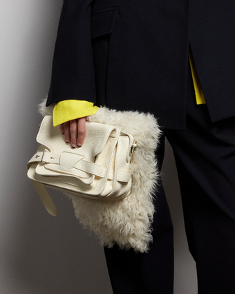 close cropped image of an off white bag against a shearling scarf held by someone with nails that have black tips, wearing a black blazer and pants with a yellow button down peeking out