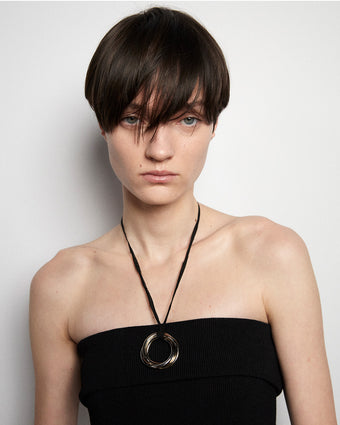 close crop of model's head and shoulders and a bit of a strapless black dress showing with a black and gold necklace, model's hair is short and hangs in front of their face