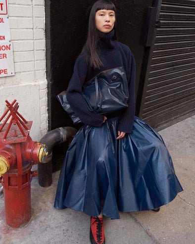 Cropped image of model sitting on the sidewalk wearing the Petra Skirt in Lacquered Cotton in Navy and the Arcadia Sneakers in Red holding the Days Carryall Bag in Navy