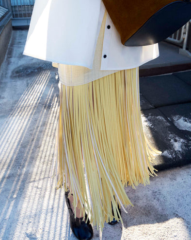 Cropped image of model standing wearing the Addy Shirt in Compact Cotton Poplin, Veda Skirt in Faux Leather Fringe in Pale Yellow Multi, and the Glove Hinge Pumps in Black holding the Brant Bucket Bag in Soft Suede