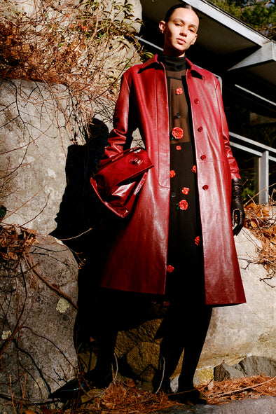 Proenza Schouler Front full length image of model wearing Nissi Coat In Semigloss Leather in Burgundy standing against rocks.