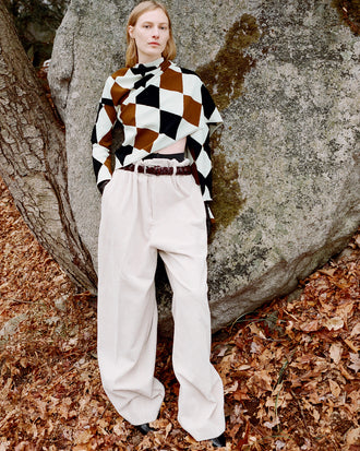 Model standing against stone boulder wearing Vanne Top in Light Green Argyle Velvet Knit, Salome Pant in Ecru Corduroy, Braid Belt in Bordeaux Croc Embossed Calf, and Uma Ankle Boots in Black Silk Calf