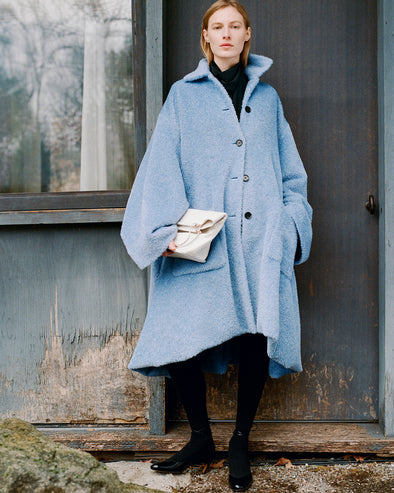 Proenza Schouler Front full length image of model standing against a wood door wearing Verretta Coat In Alpaca Coating in Sky Blue