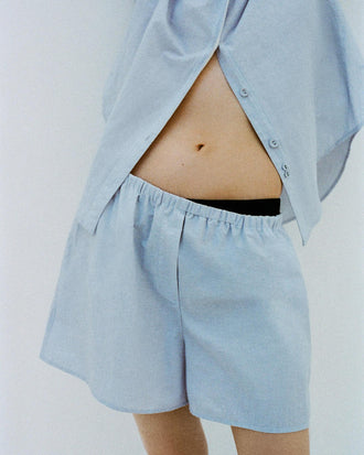 Cropped image of a model leaning against a white backdrop, wearing Caroline Shirt in Linen Shirting and Jana Short in Linen Shirting in Light Oxford Melange