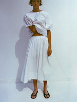 Model wearing Haleigh Top and Austin Skirt in Cotton Sateen in white, her hand looped through her top, resting against her neck