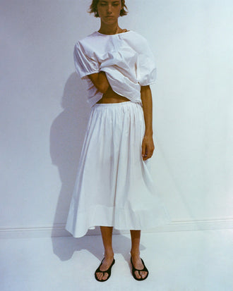 Model wearing Haleigh Top and Austin Skirt in Cotton Sateen in white, her hand looped through her top, resting against her neck