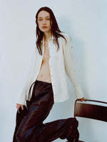 Image of model wearing Maddi Shirt in Merino Wool Twill in Bone and Magnus Pants in Lightweight Leather in Black holding onto a chair against a plain background