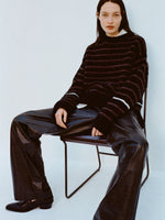 Image of model wearing Leonie Sweater in Stripe Wool in Black multi and Magnus Pants in Lightweight Leather in Black sitting on a chair against a light blue background
