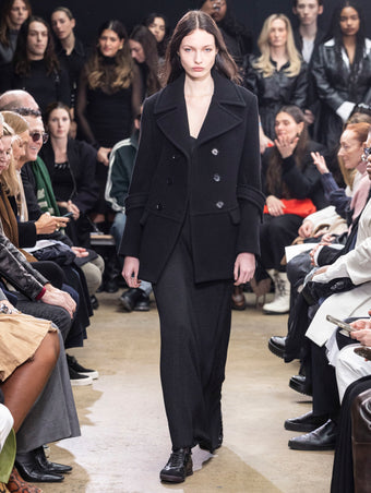Model walking in Proenza Schouler Fall Winter 2024 Runway show wearing ROWEN PEACOAT IN BLACK ECO DOUBLE FACE WOOL, BELLA DRESS IN BLACK LACQUERED KNIT, and  TRACK SNEAKERS IN BLACK NAPPA/SUEDE