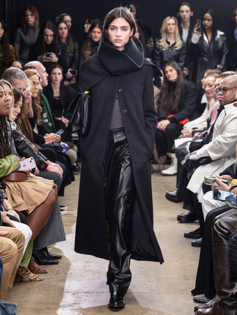 Model walking in Proenza Schouler Fall Winter 2024 Runway show wearing PHOENIX COAT IN BLACK WOOL GABARDINE, RIO TOP IN BLACK TULLE JERSEY, RORY CARGO PANT IN BLACK LEATHER, TUBE SCARF IN BLACK CASHMERE KNIT, City Messenger Bag in Black Nappa, and  BRONCO ANKLE BOOTS IN BLACK BRUSHED CALF