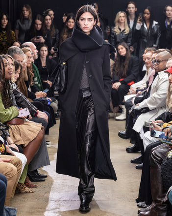 Model Walking In Proenza Schouler Fall Winter 2024 Runway Show Wearing Phoenix Coat In Black Wool Gabardine, Rio Top In Black Tulle Jersey, Rory Cargo Pant In Black Leather, Tube Scarf In Black Cashmere Knit, City Messenger Bag In Black Nappa, And  Bronco Ankle Boots In Black Brushed Calf