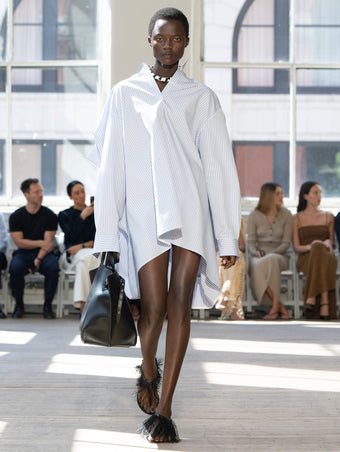 Image of model walking Proenza Schouler SS25 Runway wearing Isla Dress in Stripe Cotton Silk Poplin in Pale Blue Multi and Days Carryall in Black Smooth Calf in BLACK and Tee Feather Thong Sandal in BLACK
