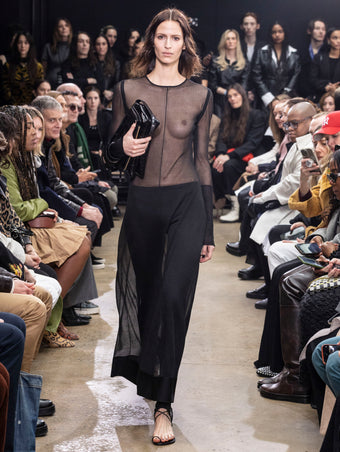 Model Walking in Proenza Schouler Fall Winter 2024 Runway Show Wearing Anneliese Dress in Black Silk Knit, Stiltsman Stirrup in Black Compact Jersey, City Bag in Black Eel, and Tee Toe Ring Sandals in Black Nappa