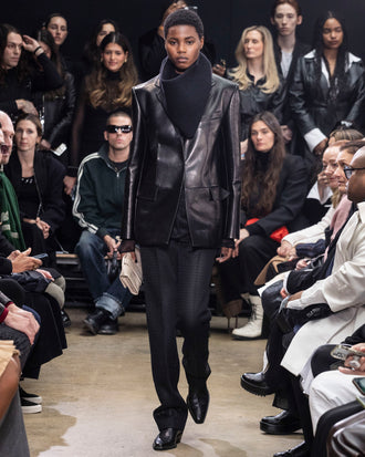 Model Walking in Proenza Schouler Fall Winter 2024 Runway Show Wearing Hayes Jacket in Black Leather, Rio Top in Black Tulle Jersey, Chase Pant in Black Technical Mesh Suiting, Tube Scarf in Black Cashmere Knit, Flip Shoulder Bag in Taupe Eel, and  Bronco Chelsea Boots in Black Box Calf