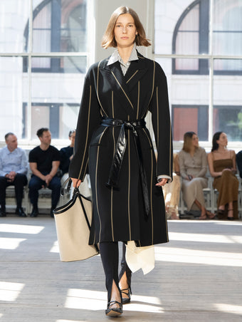 Image of model walking Proenza Schouler SS25 Runway wearing Marina Coat in Stripe Bi-Stretch Viscose in Black Multi and Hunter Shirt in Stripe Cotton Silk Poplin in Navy Multi and Zip Legging in Technical Jersey in Black and Brant Bucket Bag in Natural Linen in NATURAL / BLACK and Slash Slingback Pump in BLACK