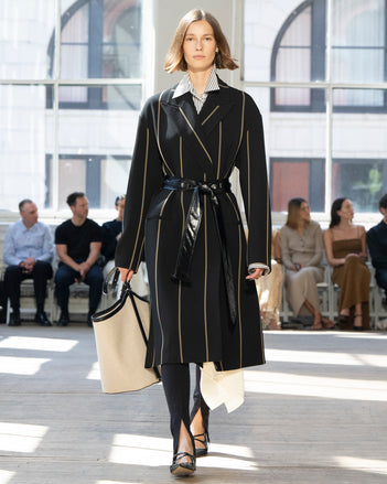 Image of model walking Proenza Schouler SS25 Runway wearing Marina Coat in Stripe Bi-Stretch Viscose in Black Multi and Hunter Shirt in Stripe Cotton Silk Poplin in Navy Multi and Zip Legging in Technical Jersey in Black and Brant Bucket Bag in Natural Linen in NATURAL / BLACK and Slash Slingback Pump in BLACK