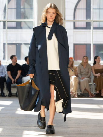 Image of model walking Proenza Schouler SS25 Runway wearing Sabina Coat in Technical Nylon Jacquard in Midnight and Renne Top in Textured Leather in Ivory and Rae Skirt in Stripe Bi-Stretch Viscose in Black Multi and Brant Bucket Bag in Black Smooth Calf in BLACK and Caribou Mule in Black