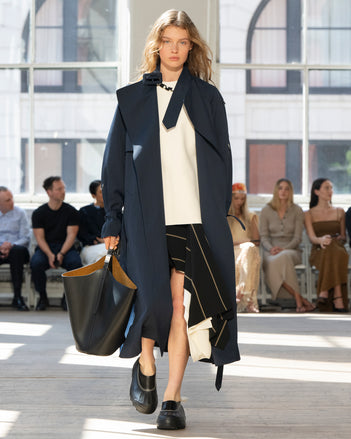 Image of model walking Proenza Schouler SS25 Runway wearing Sabina Coat in Technical Nylon Jacquard in Midnight and Renne Top in Textured Leather in Ivory and Rae Skirt in Stripe Bi-Stretch Viscose in Black Multi and Brant Bucket Bag in Black Smooth Calf in BLACK and Caribou Mule in Black