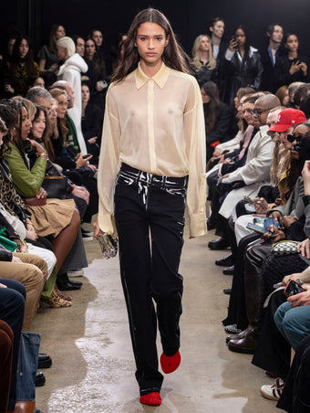 Model Walking in Proenza Schouler Fall Winter 2024 Runway Show Wearing Smithson Shirt in Resin Silk Knit, Ryman Jean in Black Garment Printed Denim, Crush Clutch in Silver Metal, Thin Belt in Black Nappa, and Tee Over the Knee Boots in Vermillion Viscose Knit