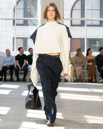 Image of model walking Proenza Schouler SS25 Runway wearing Terese Sweater in Wool Cashmere in Off White and Oliver Pant in Technical Nylon Jacquard in Midnight and Days Carryall in Black Smooth Calf in BLACK and Glove Shoe in BLACK