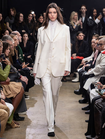 Model Walking in Proenza Schouler Fall Winter 2024 Runway Show Wearing Archer Jacket in Wax Wool Twill , Rio Top in Bone Tulle Jersey, Demi Shirt in Bone Silk Viscose, Teddy Pant in Wax Wool Twill, Silo Bag in Optic White Knotted Nappa, and Tee Over the Knee Boots in Black Viscose Knit With White Ties