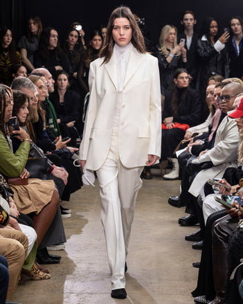 Model Walking in Proenza Schouler Fall Winter 2024 Runway Show Wearing Archer Jacket in Wax Wool Twill , Rio Top in Bone Tulle Jersey, Demi Shirt in Bone Silk Viscose, Teddy Pant in Wax Wool Twill, Silo Bag in Optic White Knotted Nappa, and Tee Over the Knee Boots in Black Viscose Knit With White Ties