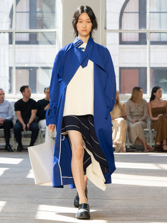 Image of model walking Proenza Schouler SS25 Runway wearing Sabina Coat in Technical Nylon Jacquard in Cobalt and Renne Top in Textured Leather in Ivory and Rae Skirt in Stripe Bi-Stretch Viscose in Navy Multi and Brant Bucket Bag in Smoke Smooth Calf in SMOKE and Caribou Mule in Black