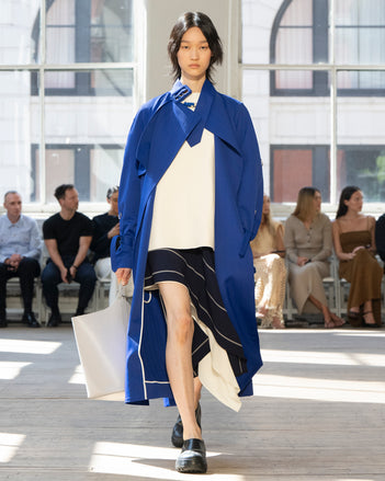 Image of model walking Proenza Schouler SS25 Runway wearing Sabina Coat in Technical Nylon Jacquard in Cobalt and Renne Top in Textured Leather in Ivory and Rae Skirt in Stripe Bi-Stretch Viscose in Navy Multi and Brant Bucket Bag in Smoke Smooth Calf in SMOKE and Caribou Mule in Black