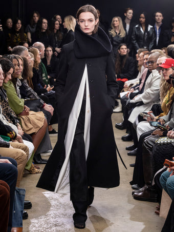 Model Walking in Proenza Schouler Fall Winter 2024 Runway Show Wearing Phoenix Coat in Black Wool Gabardine, Phoenix Coat in Ecru Wool Gabardine, Rio Top in Black Tulle Jersey, Teddy Pant in Black Double Weave Viscose Organza, Tube Scarf in Black Cashmere Knit, and Tee Over the Knee Boots in Black Viscose Knit