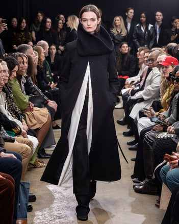 Model Walking in Proenza Schouler Fall Winter 2024 Runway Show Wearing Phoenix Coat in Black Wool Gabardine, Phoenix Coat in Ecru Wool Gabardine, Rio Top in Black Tulle Jersey, Teddy Pant in Black Double Weave Viscose Organza, Tube Scarf in Black Cashmere Knit, and Tee Over the Knee Boots in Black Viscose Knit