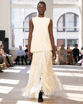 Image of model walking Proenza Schouler SS25 Runway wearing Noa Top in Textured Leather in Ivory and Effe Skirt in Compact Viscose Knit in Off White and Claudia Skirt in Silk Organza Fringe in Off White and Slash Tie Sandal in BLACK