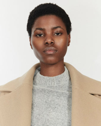 portrait image of a model looking directly at the camera wearing a light grey knit sweater and light tan coat standing against a white background