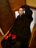 Image of model wearing Verretta Coat in Alpaca Coating in Chocolate holding Silo Clutch in Shiny Calf in Red on a white chair with wooden frame