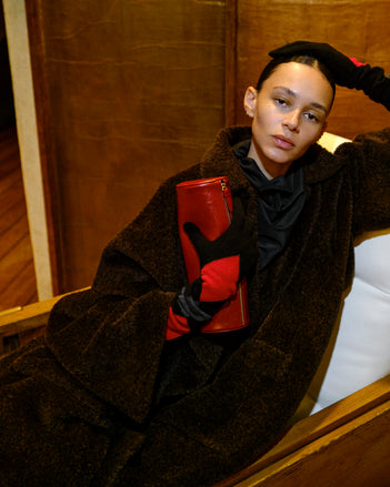 Person wearing a dark coat and red leather glove, holding the red Silo Clutch sitting on a wooden bench.
