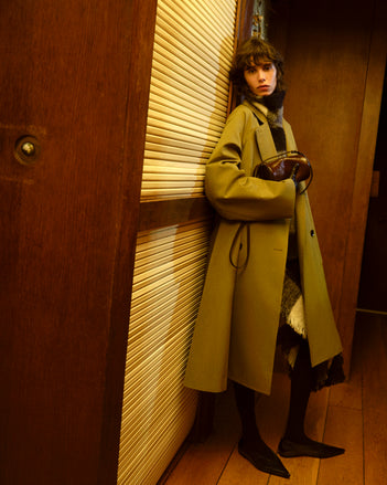 Person wearing a long green coat standing in a room with wooden walls.