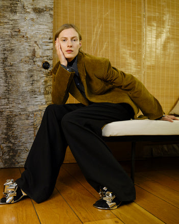 Person wearing a brown coat and black pants sitting on a chair with a textured wall background