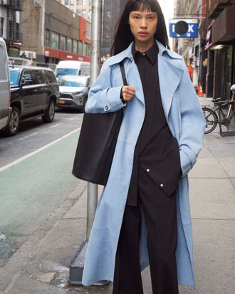 Cropped image of model standing on the sidewalk wearing the Ramona Coat in Suede in Light Blue, the Addy Shirt in Compact Poplin in Black, carrying the Brant Bucket Bag in Smooth in Black on a New York City Street
