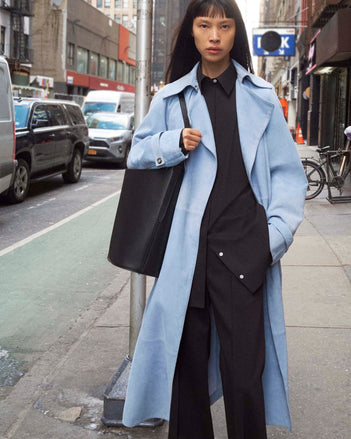 Cropped image of model standing on the sidewalk wearing the Ramona Coat in Suede in Light Blue, the Addy Shirt in Compact Poplin in Black, carrying the Brant Bucket Bag in Smooth in Black on a New York City Street