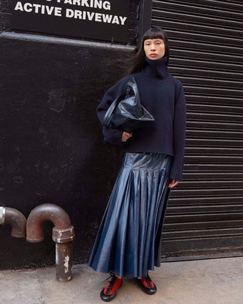Image of model standing on the sidewalk wearing the Petra Skirt in Lacquered Cotton in Navy and the Arcadia Sneakers in Red holding the Days Carryall Bag in Navy