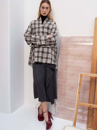 Cropped image of model standing against a white wall wearing the Bronwen Top in Crinkled Plaid in Bark Multi, Amery Skirt in Textured Suiting, and the Glove Hinge Pumps in Merlot