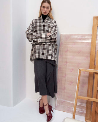 Cropped image of model standing against a white wall wearing the Bronwen Top in Crinkled Plaid in Bark Multi, Amery Skirt in Textured Suiting, and the Glove Hinge Pumps in Merlot