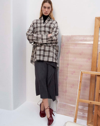 Cropped image of model standing against a white wall wearing the Bronwen Top in Crinkled Plaid in Bark Multi, Amery Skirt in Textured Suiting, and the Glove Hinge Pumps in Merlot