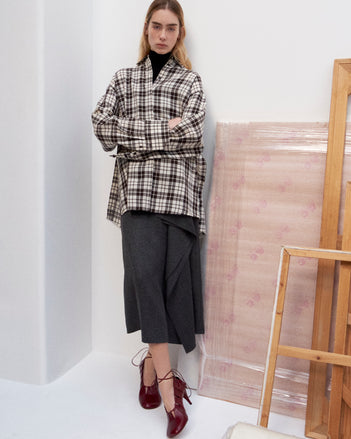 Cropped image of model standing against a white wall wearing the Bronwen Top in Crinkled Plaid in Bark Multi, Amery Skirt in Textured Suiting, and the Glove Hinge Pumps in Merlot