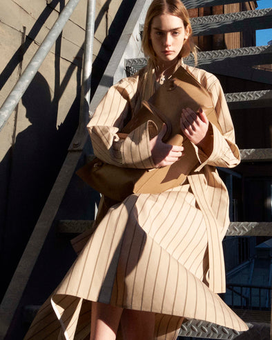 Cropped image of model sitting on a stairwell in the Phillipa Dress in Stripe Cotton Silk Poplin in Taupe Multi holding the Days Carryall in Smooth Calf in Cumin