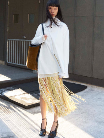 Cropped image of model standing wearing the Addy Shirt in Compact Cotton Poplin, the Veda Skirt in Faux Leather Fringe in Pale Yellow Multi, and the Glove Hinge Pumps in Black holding the Brant Bucket Bag in Soft Suede