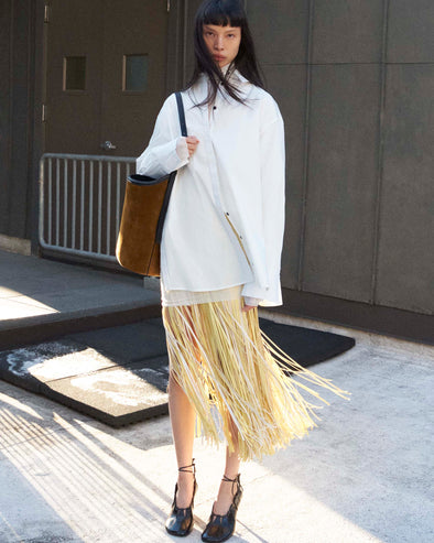 Cropped image of model standing wearing the Addy Shirt in Compact Cotton Poplin, the Veda Skirt in Faux Leather Fringe in Pale Yellow Multi, and the Glove Hinge Pumps in Black holding the Brant Bucket Bag in Soft Suede